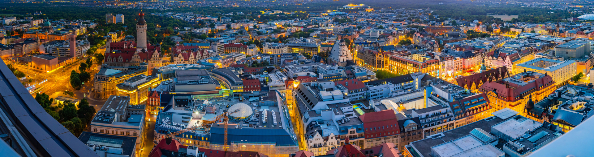 Nachtaufnahme der Skyline von Leipzig mit Blick auf das imposante City-Hochhaus, das als eines der Wahrzeichen der Stadt gilt. Die beleuchteten Straßen schlängeln sich durch das Stadtzentrum und erzeugen ein beeindruckendes Lichtspiel. Im Vordergrund ist das Gewandhaus zu erkennen, während sich daneben die futuristische Universitätskirche St. Pauli mit ihrer modernen Glasfassade erstreckt. Im Hintergrund ragen historische Gebäude und moderne Stadtstrukturen in die nächtliche Skyline, während der Himmel in einem dunklen Blau mit letzten Lichtspuren des Tages leuchtet.