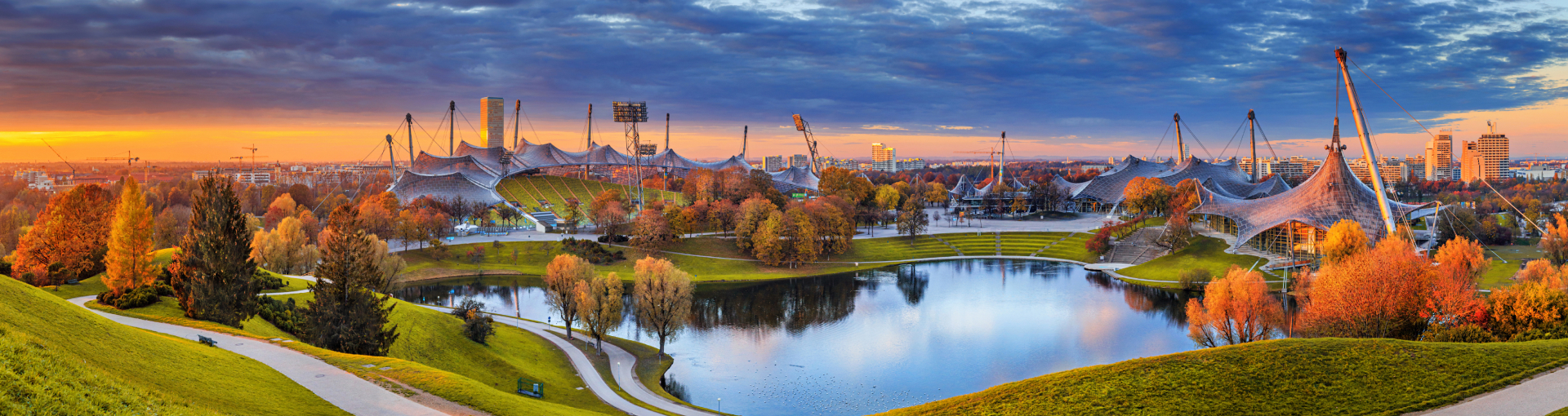 Panoramaaufnahme von München mit dem Olympiapark und dem markanten Olympiaturm im Vordergrund. Die ikonischen Zeltdachstrukturen des Olympiastadions erstrecken sich über das Gelände. Die Sonne geht hinter den Wolken unter und taucht die Szene in ein goldenes Licht. Im Hintergrund sind moderne Hochhäuser und Wohngebiete zu sehen, während die Straßen von Autos belebt sind.
