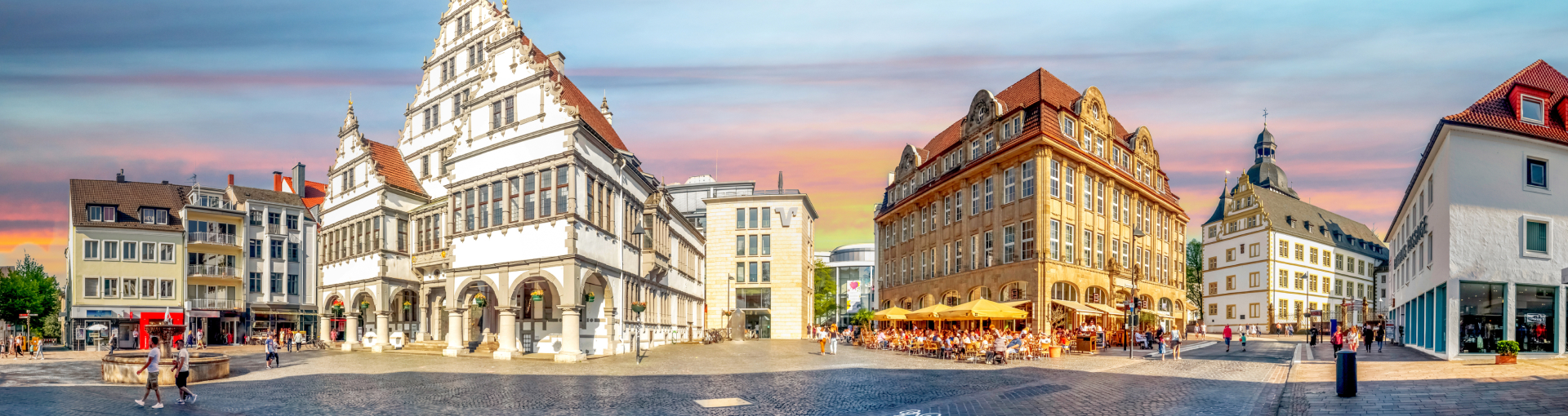 Panoramaaufnahme eines belebten Platzes in Paderborn mit historischen Gebäuden und gemütlichen Straßencafés. Im Mittelpunkt steht das prächtige Paderborner Rathaus mit seiner aufwendig verzierten Fassade im Stil der Weserrenaissance. Die umliegenden Gebäude, darunter auch das Schloss Neuhaus mit seinem markanten Turm, spiegeln die historische Architektur der Stadt wider. Der Platz ist von Menschen belebt, die einkaufen, spazieren gehen oder in Straßencafés sitzen. Ein Brunnen in der Mitte des Platzes sorgt für eine angenehme Atmosphäre, während das warme Licht des Sonnenuntergangs die Szene in sanfte Farben taucht.