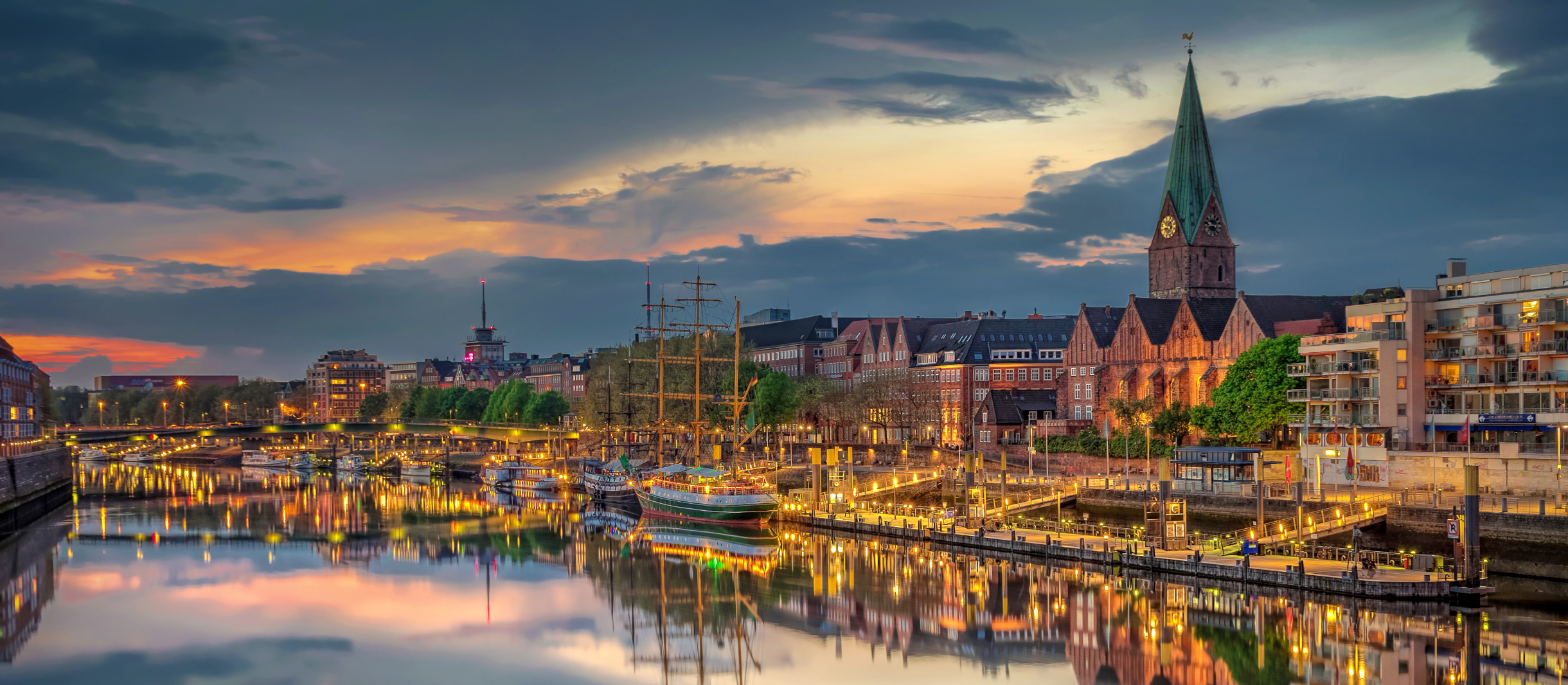 Abendliche Skyline von Bremen mit Blick auf die Weser. Die beleuchtete Uferpromenade mit historischen Gebäuden, darunter die St.-Martini-Kirche mit ihrem markanten grünen Turm, spiegelt sich im Wasser. Mehrere Segelschiffe liegen im Hafen, während der Himmel in warmen Sonnenuntergangsfarben leuchtet und sich Wolken über der Stadt verteilen.