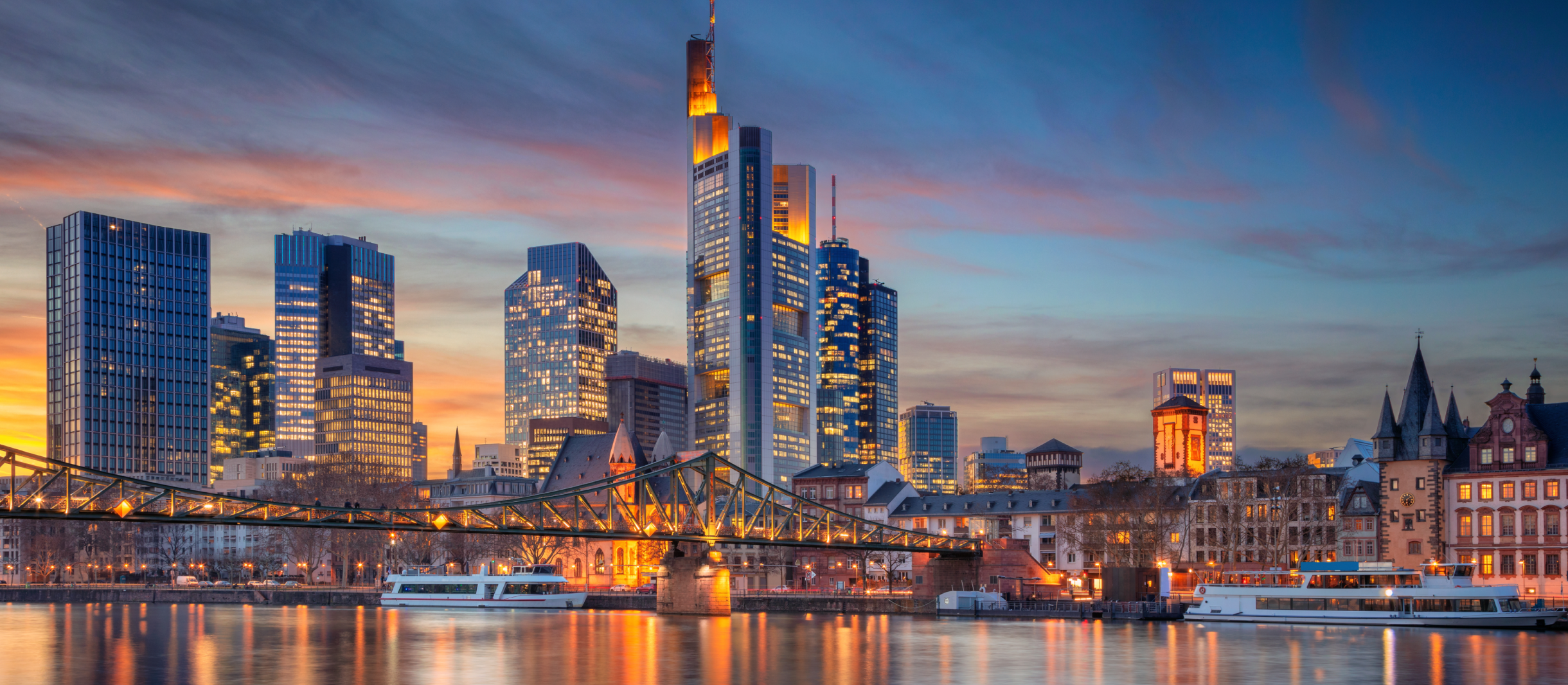 Abendliche Skyline von Frankfurt am Main mit leuchtenden Wolkenkratzern und dem charakteristischen Commerzbank Tower. Im Vordergrund fließt der Main, über den eine Brücke mit beleuchteten Geländern führt. Die untergehende Sonne taucht den Himmel in warme Orange- und Blautöne, während die Lichter der Stadt das Wasser reflektieren. Moderne Hochhäuser kontrastieren mit historischen Gebäuden am Flussufer.