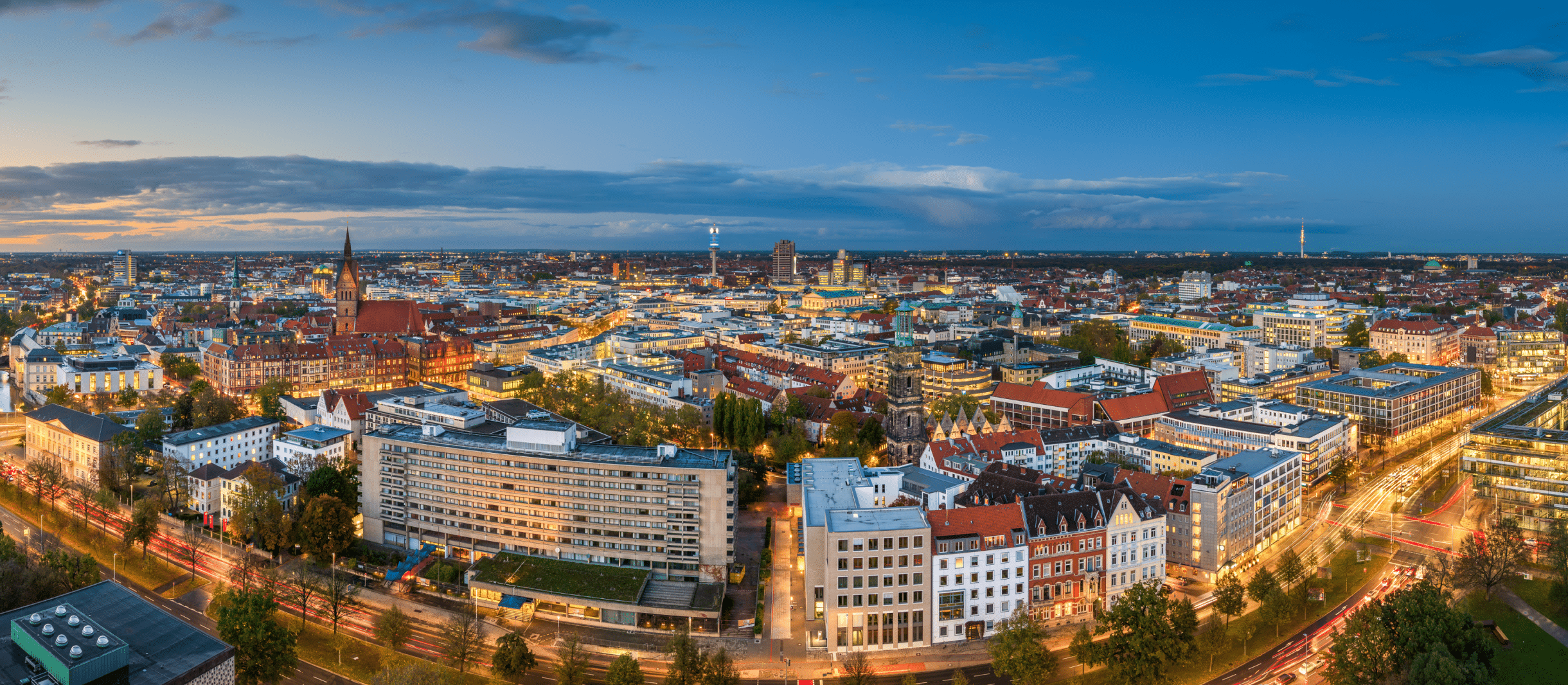 Panoramaansicht der Stadt Hannover bei Abenddämmerung mit beleuchteten Straßen, modernen Gebäuden und historischen Bauwerken. Im Zentrum der Stadt ragt die Marktkirche mit ihrem markanten roten Backsteinturm empor. Die Stadt ist von grünen Bäumen und zahlreichen Straßen mit Lichtspuren vorbeifahrender Autos durchzogen. Im Hintergrund erstreckt sich der Himmel mit blauen und orangefarbenen Tönen, während sich das Stadtleben in warmem Licht entfaltet.