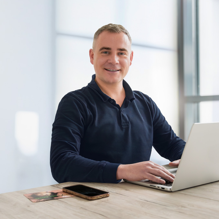 Ein freundlicher Mann mittleren Alters mit kurzen, hellen Haaren sitzt an einem Schreibtisch und arbeitet an einem Laptop. Er trägt ein dunkles, langärmliges Polohemd und lächelt in die Kamera. Neben ihm liegt ein Smartphone auf dem Tisch. Der helle, moderne Büroraum im Hintergrund ist lichtdurchflutet.