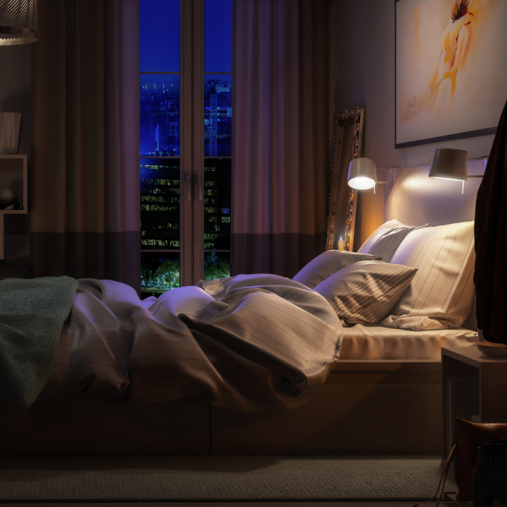 Ein gemütliches Schlafzimmer bei Nacht mit gedämpfter Beleuchtung. Das Bett ist leicht unordentlich, mit weichen Kissen und einer Decke, die teilweise zurückgeschlagen ist. Durch das Fenster ist eine nächtliche Stadtlandschaft mit beleuchteten Gebäuden zu sehen. Eine warme Leselampe am Bett sorgt für eine entspannte Atmosphäre.