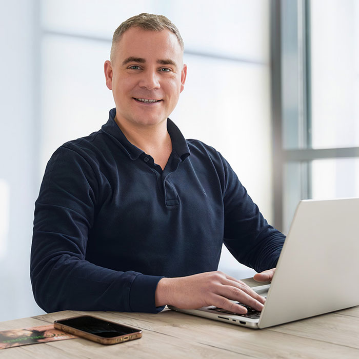 Ein freundlicher Mann mittleren Alters mit kurzen, hellen Haaren sitzt an einem Schreibtisch und arbeitet an einem Laptop. Er trägt ein dunkles, langärmliges Polohemd und lächelt in die Kamera. Neben ihm liegt ein Smartphone auf dem Tisch. Der helle, moderne Büroraum im Hintergrund ist lichtdurchflutet.