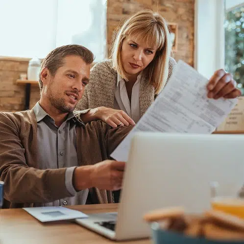 Dynamische Stromtarife im Vergleich: Paar prüft Angebote zu Hause Ein Mann in einem braunen Cardigan und eine Frau in einer beigen Strickjacke sitzen gemeinsam an einem Holztisch. Der Mann hält ein Dokument in der Hand, während die Frau sich zu ihm neigt und darauf sowie auf einen aufgeklappten Laptop blickt. Im Vordergrund steht eine Schale mit Gebäck.