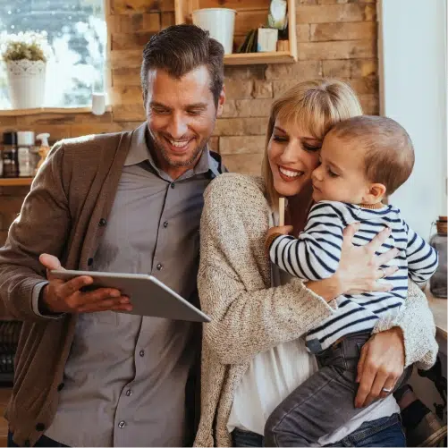 Dynamische Stromtarife für Familien: Eltern prüfen Preise am Tablet Ein Mann in einem braunen Cardigan hält ein Tablet in der Hand und zeigt den Bildschirm einer Frau, die neben ihm steht und ein Kleinkind im gestreiften Langarmshirt auf dem Arm trägt. Alle drei blicken auf das Tablet und die Erwachsenen lächeln.
